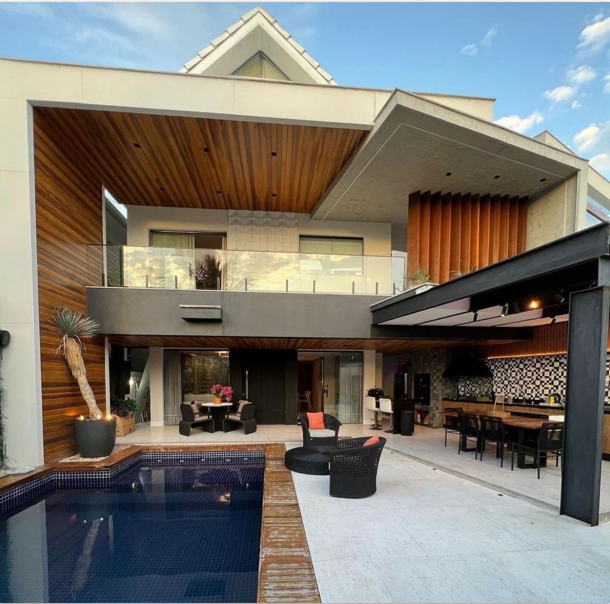 Área externa com piscina, deck de madeira, varanda gourmet coberta com mesa de jantar e poltronas, e fachada com revestimento em madeira.