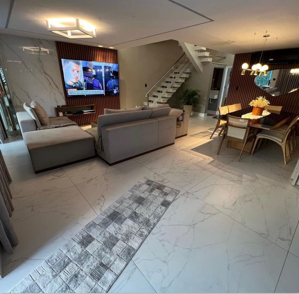 Sala de estar e jantar integrada com piso de porcelanato, painel ripado na TV e escada com degraus suspensos.