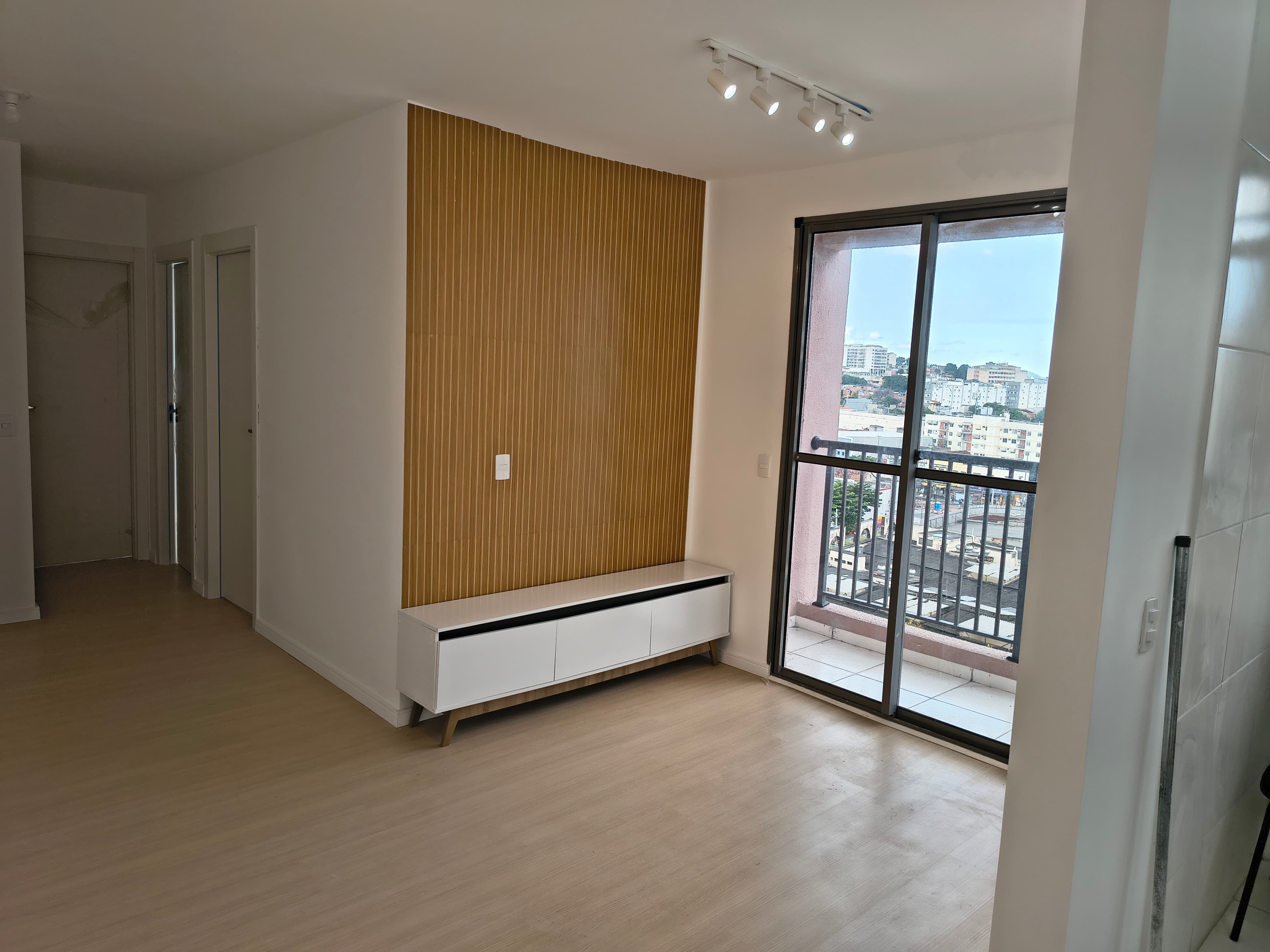 Sala com piso laminado, painel ripado em tom amadeirado e rack branco, com acesso à varanda.