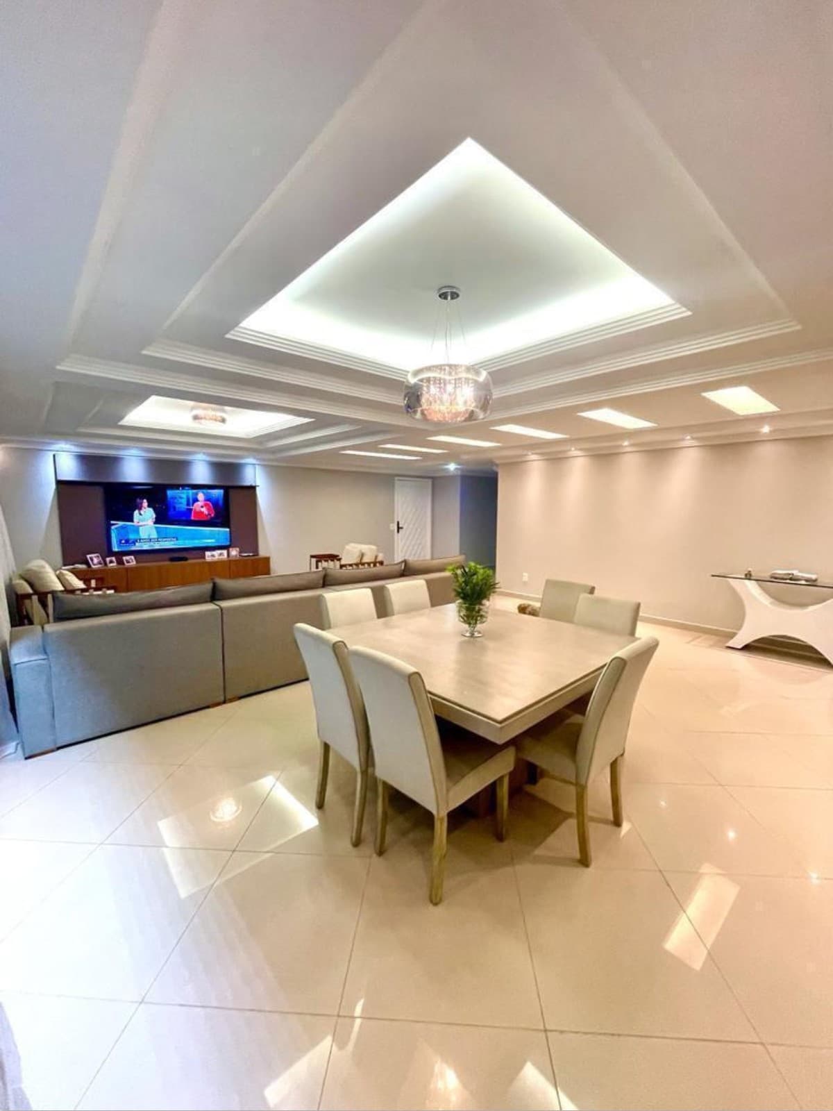 Sala de estar e jantar integrada com piso claro, teto rebaixado com iluminação embutida e lustre central.